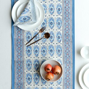 Peut inclure: Une nappe bleue et blanche à motifs avec une serviette assortie et des assiettes blanches. Le chemin de table présente un motif floral et est associé à des couverts en bronze et à un bol de grenades.