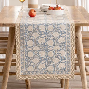 May include: A blue and beige floral patterned table runner. The runner features a repeating floral design in beige against a blue background. The edges have a striped border in beige. The runner is placed on a wooden table, with pomegranates and dishes visible.