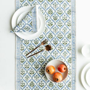 May include: A white tablecloth with a blue and green floral pattern. A white plate with a folded napkin, a bowl of pomegranates, and bronze cutlery are arranged on the table runner. The runner has a border with a repeating design.