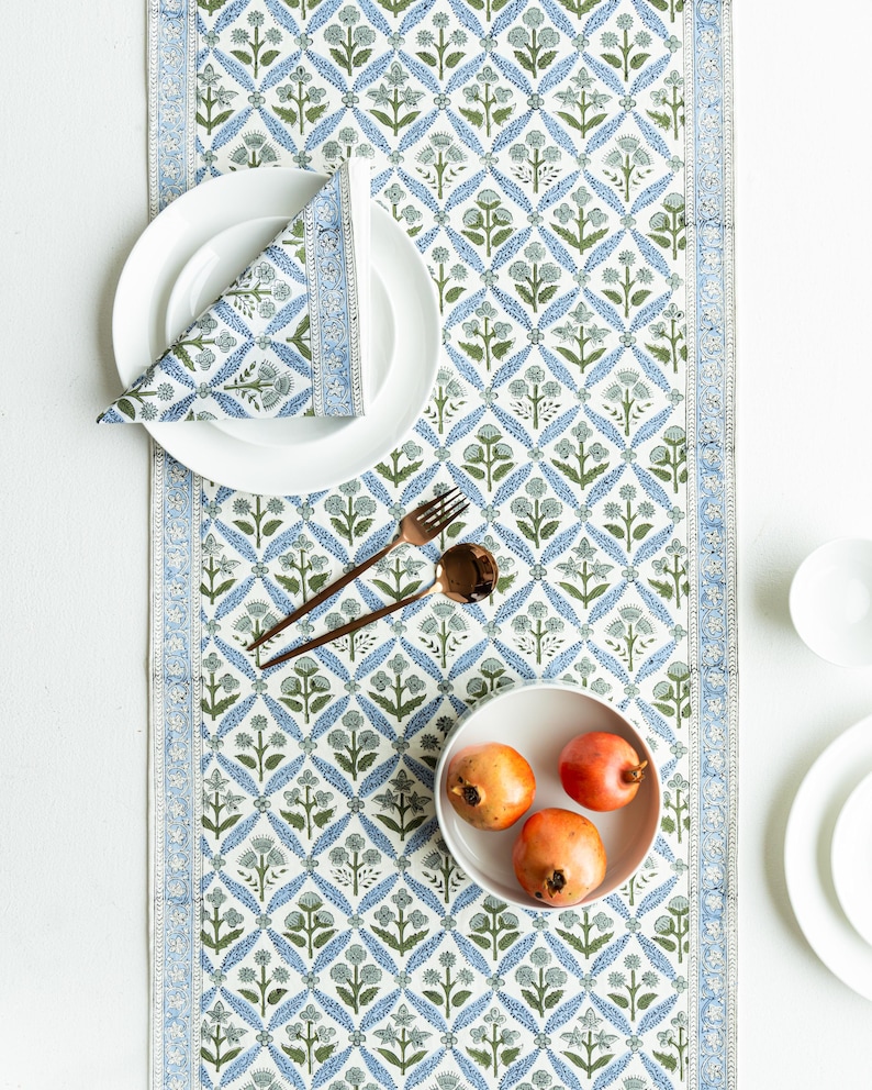 May include: A white tablecloth with a blue and green floral pattern. A white plate with a folded napkin, a bowl of pomegranates, and bronze cutlery are arranged on the table runner. The runner has a border with a repeating design.