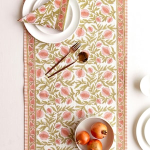 May include: A white tablecloth featuring a pattern of pink pomegranates and green foliage. A matching napkin rests on a white plate, with gold cutlery. A bowl of pomegranates is also visible.