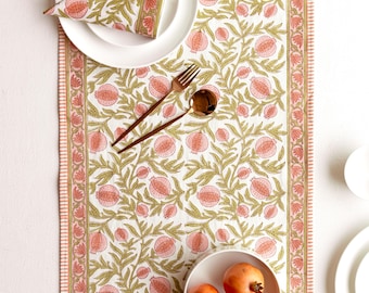 May include: A white tablecloth with a repeating pattern of pink pomegranates and green leaves. A matching napkin sits on a white plate, with gold-colored silverware. A bowl of pomegranates is also present.