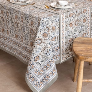 May include: A floral patterned tablecloth in shades of gray, blue, and brown, with matching placemats and napkins. The tablecloth features a detailed border design and is set on a wooden table. The overall theme is home decor.