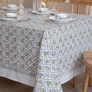 以下が含まれることがあります： 青と緑の花柄の白いテーブルクロスが、食事のためにセットされたテーブルを覆っています。 テーブルクロスは、花のアクセントが付いたダイヤモンド型のデザインです。 テーブルには、白いお皿、ボウル、金色のカトラリーがセットされています。