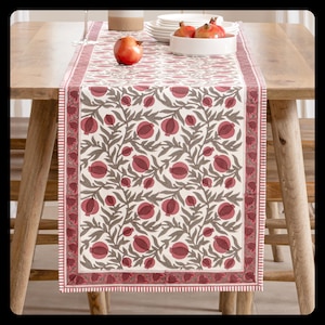 Pode incluir: Um caminho de mesa retangular com fundo creme e um padrão repetido de romãs vermelhas e folhas cinzentas. A borda é vermelha lisa com uma fina faixa branca. O caminho de mesa está sobre uma mesa de madeira.