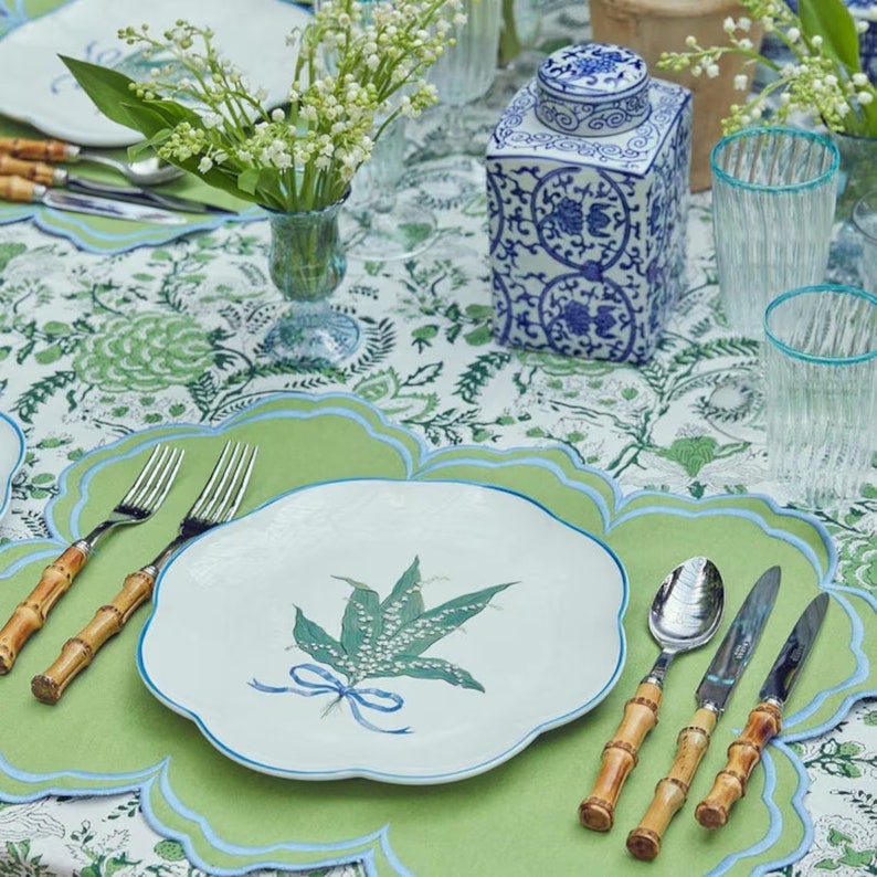 May include: A white porcelain plate with a blue rim and a green lily of the valley design. The plate is set on a green placemat with a blue scalloped edge. Two bamboo handled forks and a bamboo handled spoon are on the placemat.