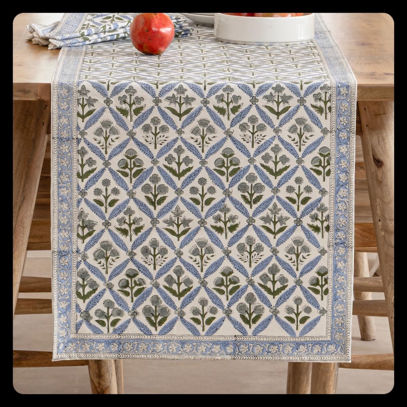 May include: A rectangular table runner with a repeating floral pattern in blue, green, and gray on a cream background. The runner has a blue border with a detailed design. A red pomegranate and a white bowl are visible on the table.