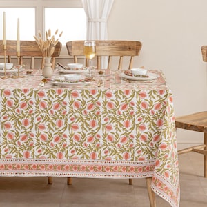Op de afbeelding: Een witte tafelkleed met een herhalend patroon van roze bloemen en groene bladeren. De tafel is gedekt met witte borden, gouden bestek en een wijnglas. Het tafelkleed heeft een decoratieve rand.