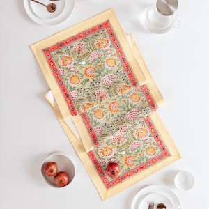 Pode incluir: Dois caminhos de mesa com padrão floral com fundo verde claro, flores laranja e rosa e borda listrada amarela. Os caminhos de mesa estão sobre uma superfície branca, com pratos, tigelas e talheres brancos. Três romãs estão em uma tigela branca.