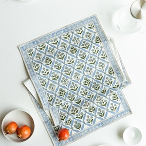 May include: Two white cotton placemats with a blue and green floral pattern. The placemats have a blue border and are placed on a white surface. The image also includes white plates, a bowl, and pomegranates.