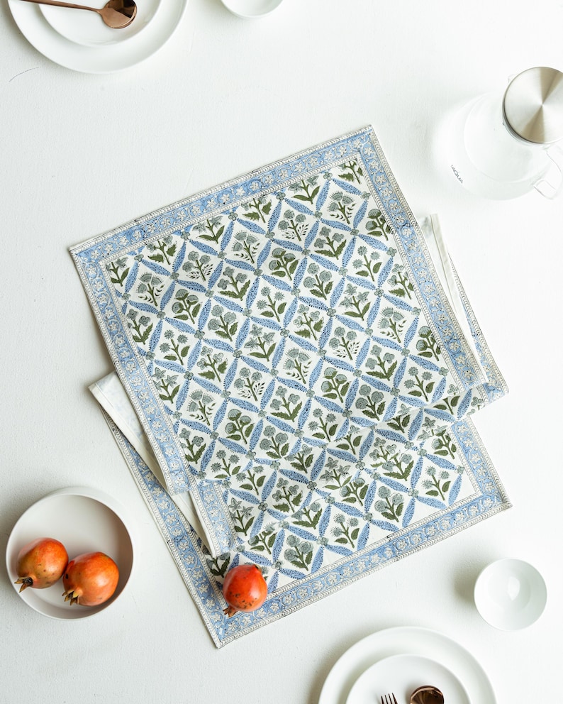 May include: Two white cotton placemats with a blue and green floral pattern. The placemats have a blue border and are placed on a white surface. The image also includes white plates, a bowl, and pomegranates.
