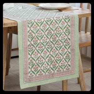 Puede incluir: Un camino de mesa floral con un patrón repetido de flores rosas y hojas verdes sobre un fondo blanco. El camino de mesa tiene un borde a rayas verdes y blancas y se muestra sobre una mesa de madera. Este camino de mesa decorativo es ideal para cualquier mesa.