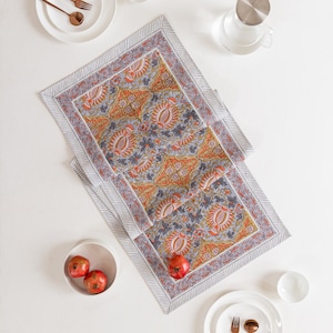 Peut inclure: Chemin de table rectangulaire avec un motif floral orange, gris et bleu. Autour du chemin de table, on trouve des assiettes blanches, des bols et des couverts cuivrés. Une carafe en verre et des grenades sont également présentes.