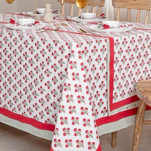 Puede incluir: Mantel blanco con un patrón repetido de diseños florales rojos. El mantel tiene un borde rojo y un ribete a rayas verdes y blancas. La mesa está puesta con platos blancos, servilletas y cubiertos dorados.