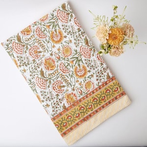 Puede incluir: Un mantel blanco con un estampado floral en naranja, amarillo y verde. El borde presenta una banda amarilla con un diseño floral y un borde a rayas. Un ramo de flores naranjas y melocotón está cerca.