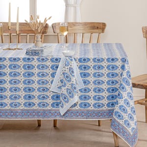 May include: A rectangular tablecloth with a repeating blue eye pattern on a white background. A matching napkin is draped over the table. The tablecloth has a decorative border with red and blue accents. The table is set with glasses, candles, and a vase.