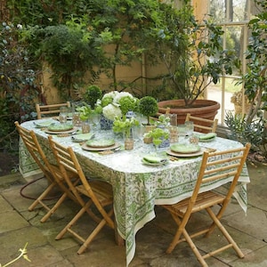 May include: A green and white patterned tablecloth covers a wooden table set for a dinner party.  The table is set with white plates, green napkins, and clear glasses.  There are white flowers in a vase in the center of the table.  The table is surrounded by six wooden chairs.