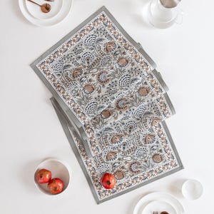 Puede incluir: Un camino de mesa gris con estampado floral y borde detallado. El camino de mesa está colocado sobre una superficie blanca, junto con platos, cuencos y utensilios de color cobre. También hay granadas rojas y una jarra de agua de cristal.