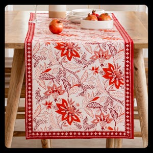 Puede incluir: Un camino de mesa rectangular con fondo blanco y un estampado floral repetido en tonos rojos. El camino de mesa tiene un borde rojo liso con un borde decorativo. Granadas y platos blancos sobre la mesa de madera.