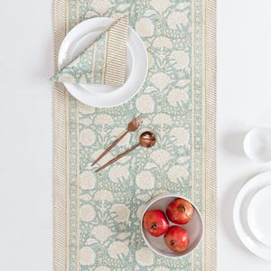 Puede incluir: Una mesa con un camino de mesa floral en verde azulado y crema, con servilleta a juego y platos blancos. Cubiertos de color cobre y un cuenco con granadas completan el conjunto, creando una elegante escena de comedor.