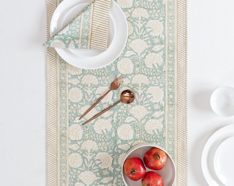 May include: A table setting featuring a floral patterned table runner in teal and cream, with matching napkin and white plates. Copper-colored flatware and a bowl of pomegranates complete the arrangement, creating an elegant dining scene.