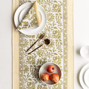 Pode incluir: Uma mesa posta com um prato branco, uma toalha de mesa dobrada e talheres de bronze. Uma tigela contém romãs. O caminho de mesa e a toalha de mesa apresentam um padrão floral verde e branco, com bordas listradas amarelas.