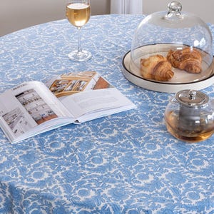 Puede incluir: Una mesa redonda cubierta con un mantel azul con estampado floral. Una copa de vino blanco, un libro, una tetera de cristal y un plato con cruasanes bajo una cúpula de cristal están sobre la mesa. El mantel es azul claro y blanco.