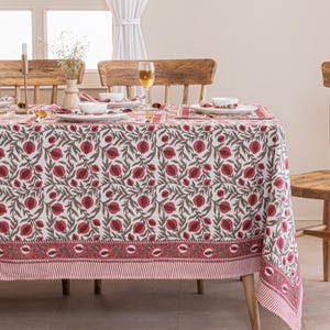 May include: A white tablecloth with a repeating pattern of red pomegranates and brown leaves. The tablecloth is set on a wooden table with matching wooden chairs. The border of the tablecloth has a red and white striped design.
