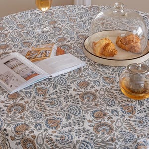 May include: A round tablecloth featuring a floral pattern in blue, beige, and white. A glass of wine, a book, a glass dome with croissants, and a teapot are on the table. The tablecloth has a detailed botanical design.