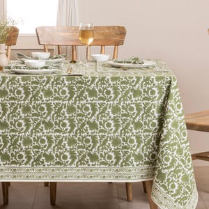 May include: A dining table is covered with a green and white floral tablecloth. The table is set with white plates, bowls, and napkins. A glass of wine sits on the table, and wooden chairs surround it.