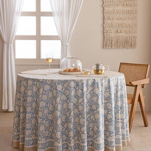 Pu&ograve; includere: Una tovaglia rotonda con un motivo floreale blu e bianco, apparecchiata per un pasto. Il tavolo &egrave; apparecchiato con una campana di vetro, una teiera e un bicchiere di vino. Sono visibili anche una sedia in vimini e un arazzo.