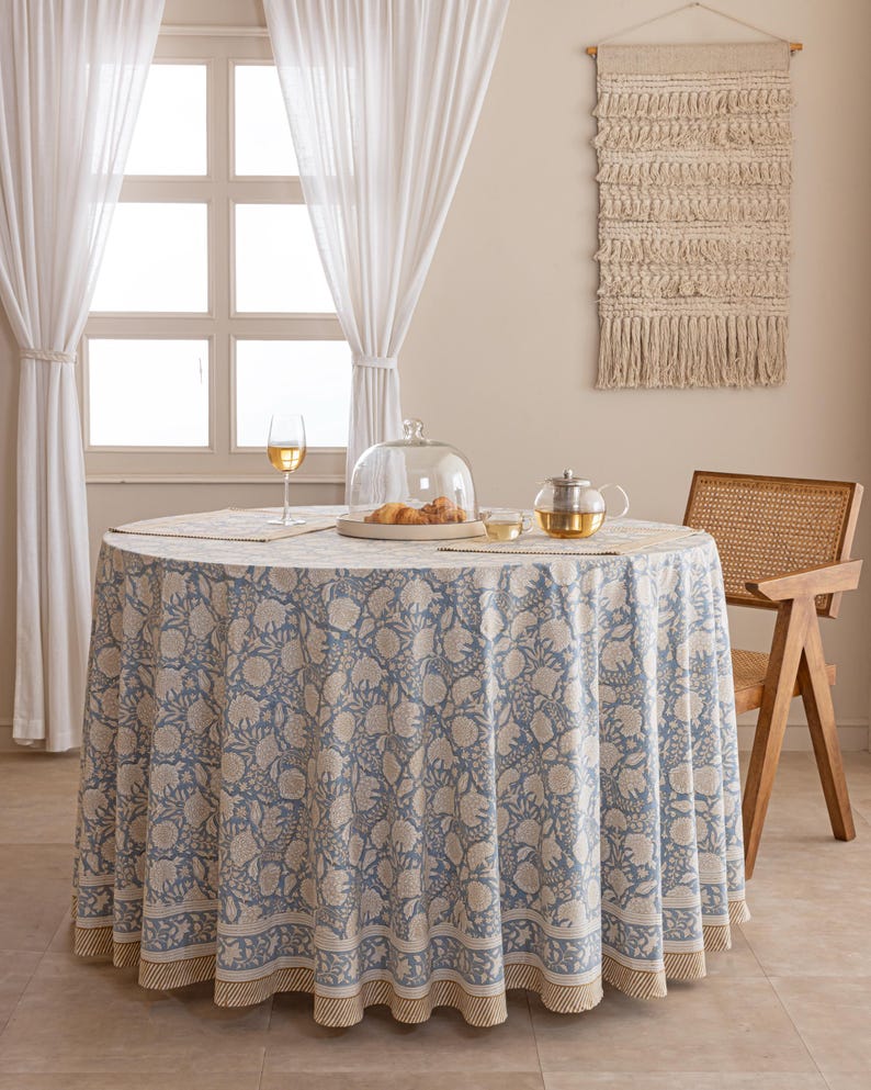 Pu&ograve; includere: Una tovaglia rotonda con un motivo floreale blu e bianco, apparecchiata per un pasto. Il tavolo &egrave; apparecchiato con una campana di vetro, una teiera e un bicchiere di vino. Sono visibili anche una sedia in vimini e un arazzo.