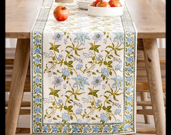 May include: A rectangular table runner with a floral pattern of blue flowers and green leaves on a cream background. The runner is placed on a wooden table, with pomegranates and white dishes visible.