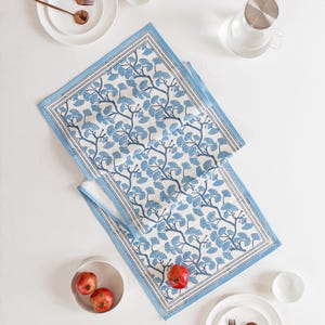 Puede incluir: Dos caminos de mesa estampados en azul y blanco con un diseño botánico. Los caminos de mesa están rodeados de platos blancos, cuencos y tenedores y cucharas de color cobre. También hay manzanas rojas y una jarra de agua de cristal.