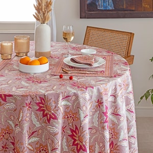 May include: A round table set for dining, featuring a floral patterned tablecloth in shades of pink, red, and cream. The table is set with plates, napkins, and silverware. A vase with dried flowers, candles, and a bowl of oranges complete the setting.
