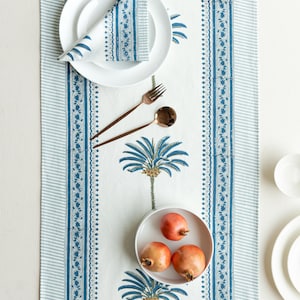 May include: A white table runner with a blue palm tree design and striped borders. The runner is set with white plates, a folded napkin, copper-colored silverware, and a small bowl with three pomegranates.