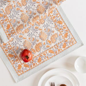 Könnte beinhalten: Ein cremefarbener Tischläufer mit orangefarbenen und grauen Blumenmustern. Der Läufer hat einen gestreiften Rand in Hellblau und Weiß. Ein roter Granatapfel, ein weißer Teller und eine Schüssel sind ebenfalls zu sehen.
