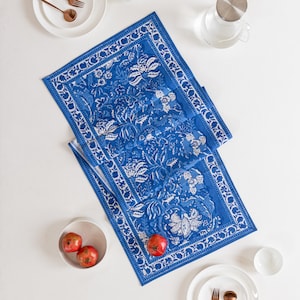 Puede incluir: Un camino de mesa azul y blanco con estampado floral. El camino de mesa tiene un diseño floral detallado y un borde blanco. La imagen también incluye platos blancos, cuencos, utensilios de color cobre, granadas y una jarra de cristal.