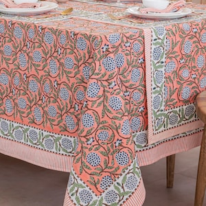 May include: A coral-colored tablecloth with a floral pattern featuring blue and green accents. The design includes stylized flowers and leaves, with a striped border. The tablecloth is set on a wooden table, ready for a meal.