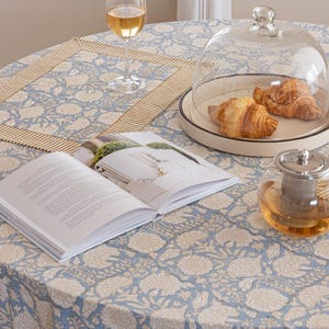Pu&ograve; includere: Una tavola apparecchiata per la colazione con una tovaglia rotonda a fantasia floreale blu. Include un bicchiere di vino, un libro, una campana di vetro con croissant e una teiera. &Egrave; presente anche un sottopiatto abbinato.