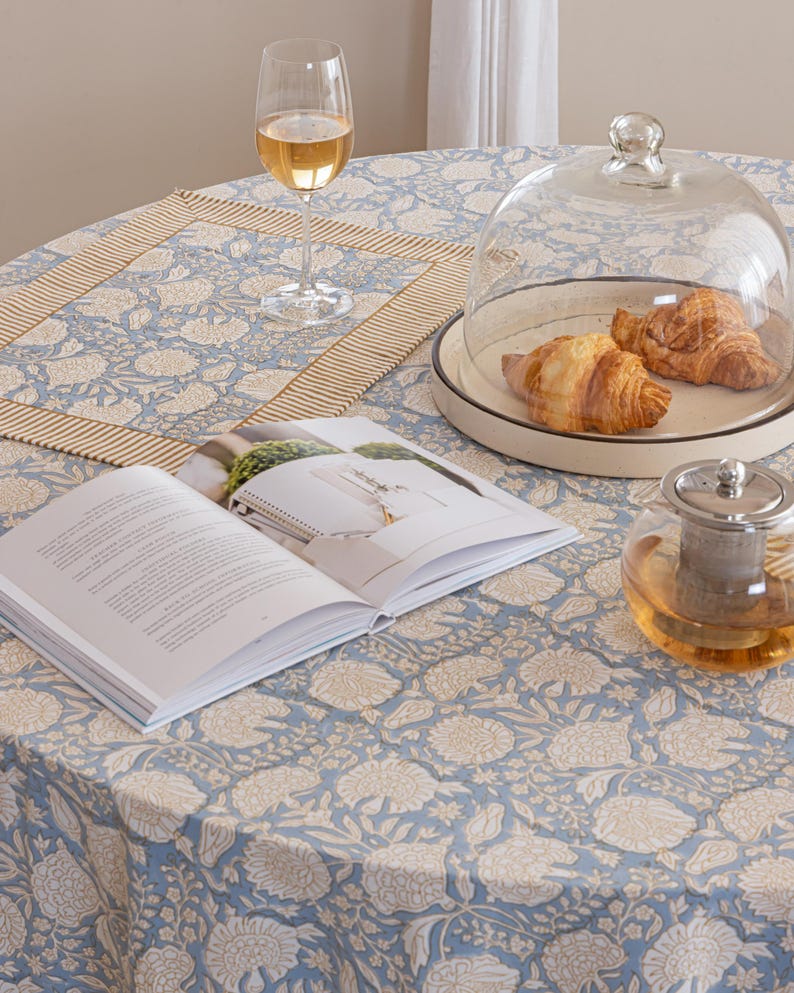 Pu&ograve; includere: Una tavola apparecchiata per la colazione con una tovaglia rotonda a fantasia floreale blu. Include un bicchiere di vino, un libro, una campana di vetro con croissant e una teiera. &Egrave; presente anche un sottopiatto abbinato.