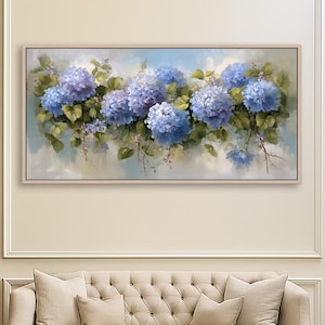 Puede incluir: Pintura floral enmarcada con vibrantes flores de hortensias azules con hojas verdes sobre un fondo suave azul claro y blanco. La obra de arte se exhibe sobre un sofá beige en un espacio interior bien iluminado.