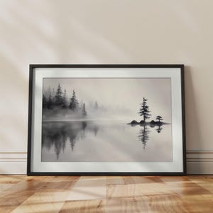 May include: A framed black and white landscape print featuring a misty lake scene with a dense forest on the left and a small island with two evergreen trees on the right. The calm water reflects the trees and fog in a soft, monochromatic tone.