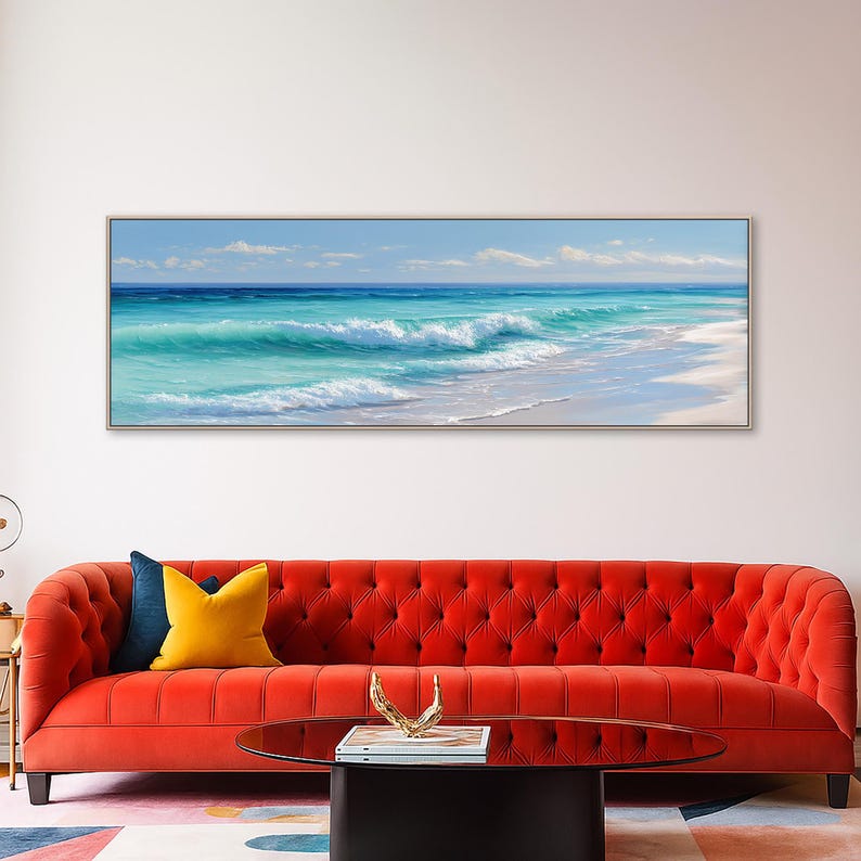 May include: A framed rectangular canvas art piece depicting a coastal scene. The artwork showcases a vibrant turquoise ocean with white-capped waves crashing onto a sandy beach under a clear blue sky. The artwork is displayed above a red sofa.