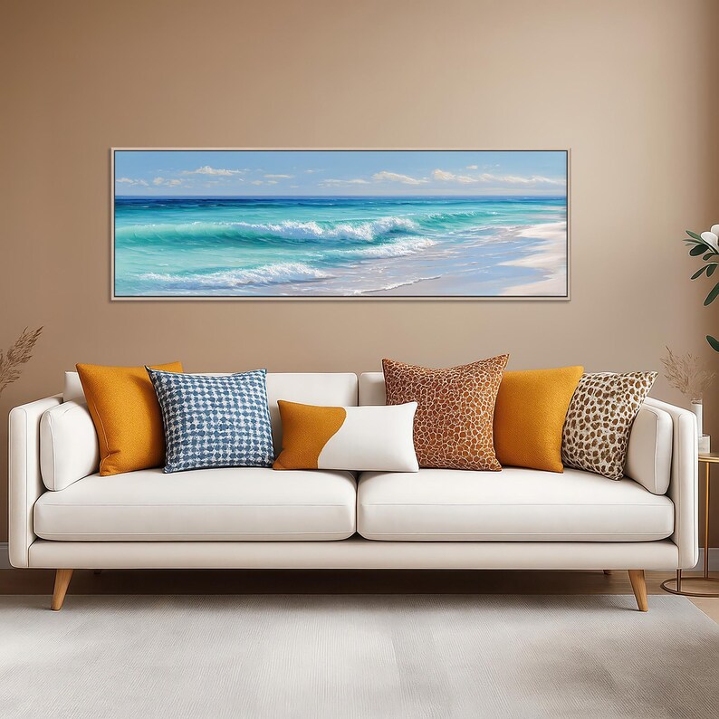 May include: A framed canvas print of a beach scene with turquoise water and white waves. The artwork hangs above a white sofa with orange, blue, and patterned throw pillows. The room has a neutral color palette.