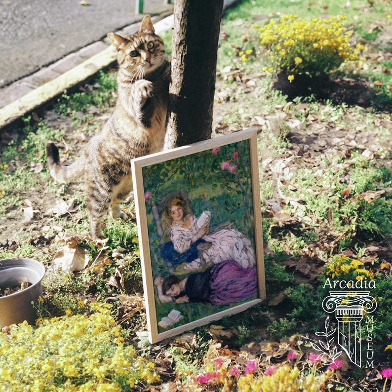 May include: A brown tabby cat stands beside a framed painting of two figures in a garden, one reading a book. The painting features a person in a white dress and another in a dark dress, surrounded by green foliage and pink flowers. The text Arcadia Museum is visible.