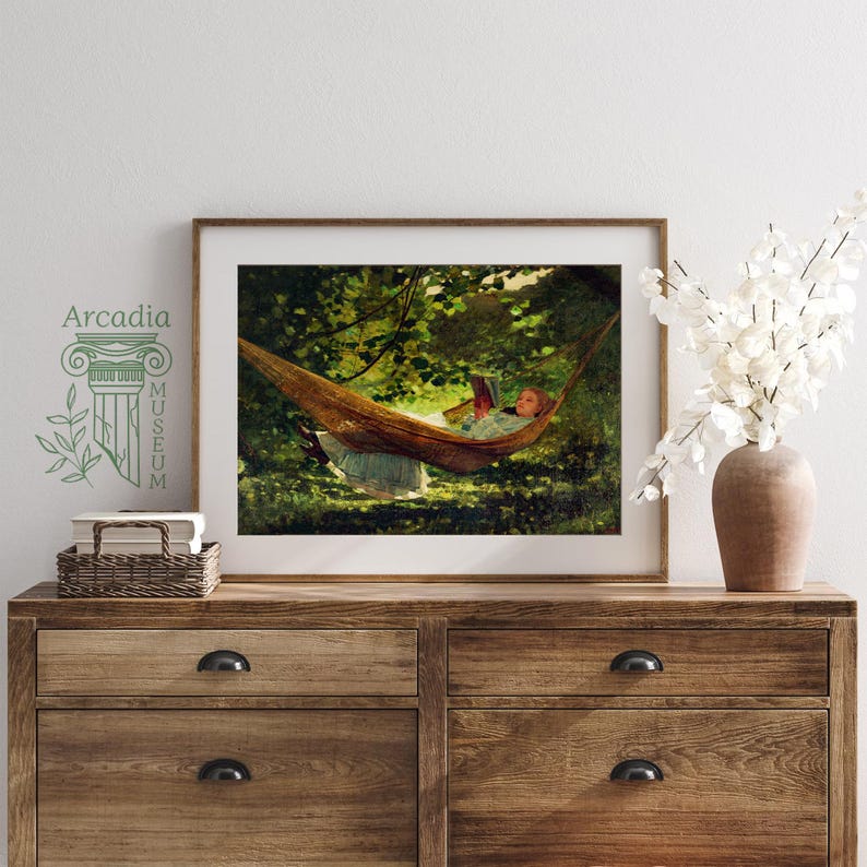 May include: A framed art print depicting a person reading in a hammock suspended between trees, displayed on a wooden dresser next to a woven basket and a vase of white flowers. The text Arcadia Museum is printed on the wall to the left.