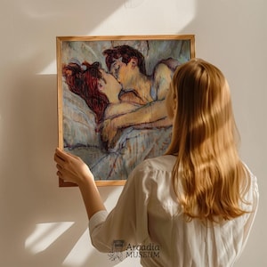 May include: A square framed art print featuring an impressionistic painting of two figures embracing and kissing while lying in bed. The artwork uses a palette of warm skin tones, deep reds, and soft blues, all enclosed within a simple light wood frame.