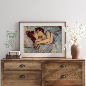 May include: A framed art print depicting a couple embracing and kissing while lying in bed, rendered in a painterly style with soft blue, tan, and red tones. The print is displayed in a wooden frame on a wooden dresser next to a vase of white flowers.