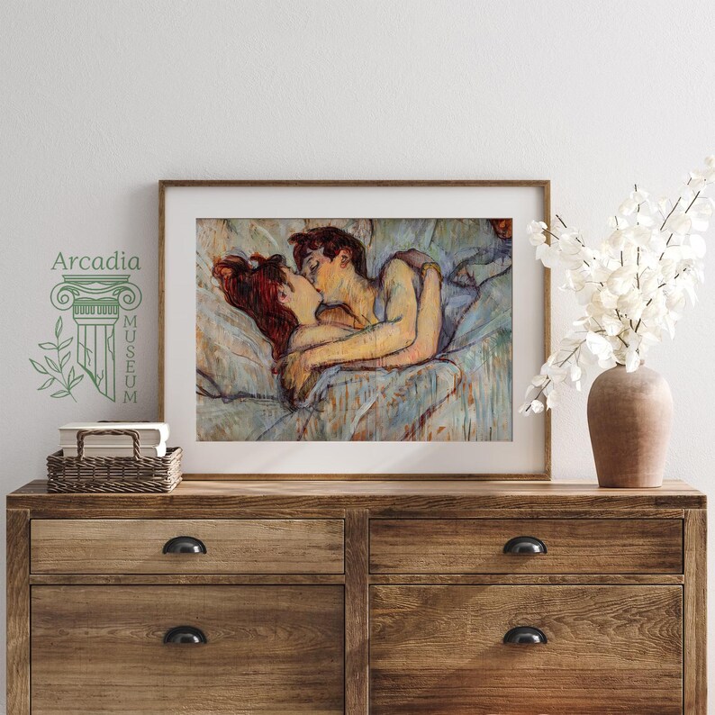 May include: A framed art print depicting a couple embracing and kissing while lying in bed, rendered in a painterly style with soft blue, tan, and red tones. The print is displayed in a wooden frame on a wooden dresser next to a vase of white flowers.